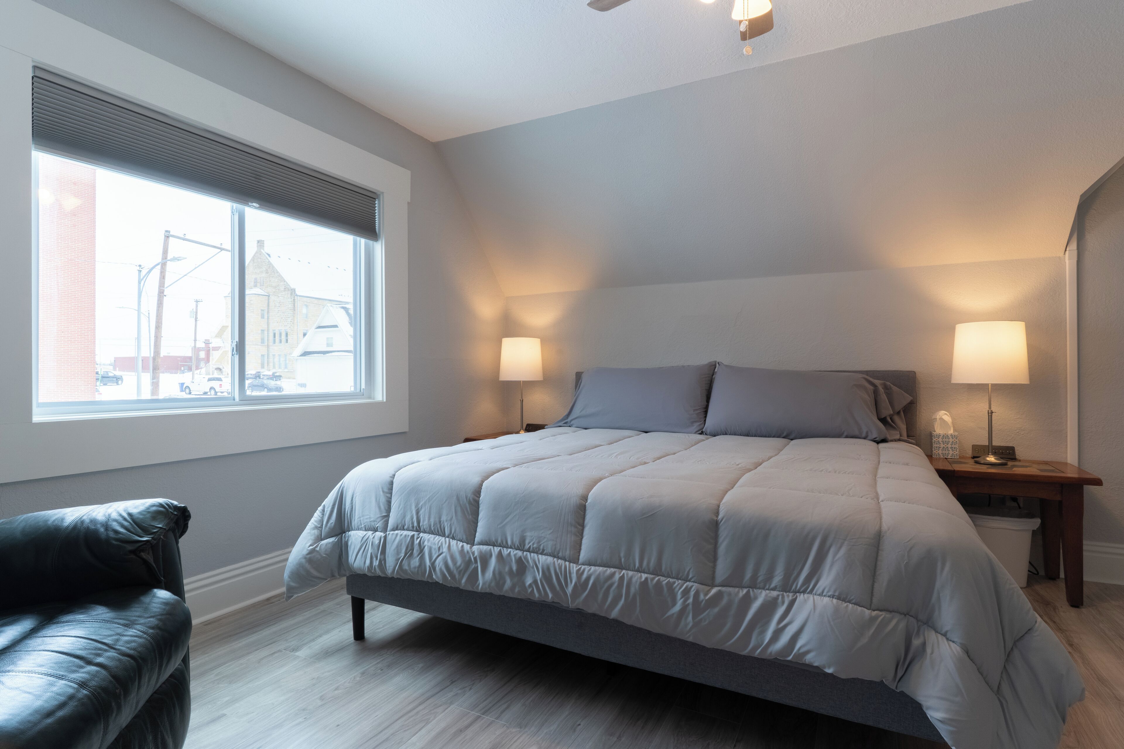A modern bedroom with a large bed, two bedside lamps, a window with blinds, and a leather chair.