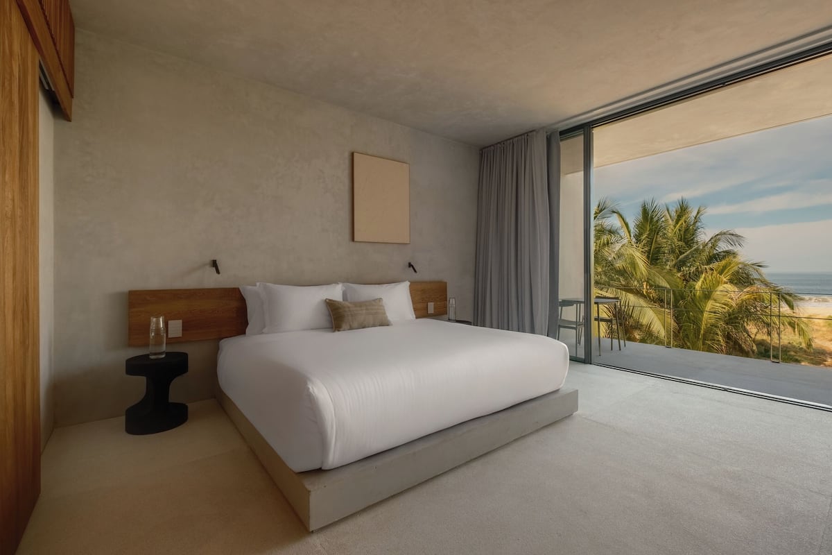 A modern hotel room with a large bed, a small bedside table, and a view of the beach.