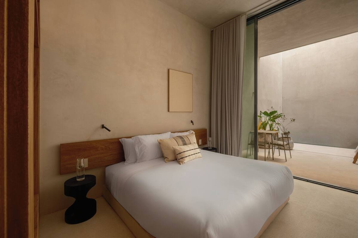 A modern hotel room with a large bed, a bedside table with a glass of water, a small table with a plant, and a view of the outside balcony.