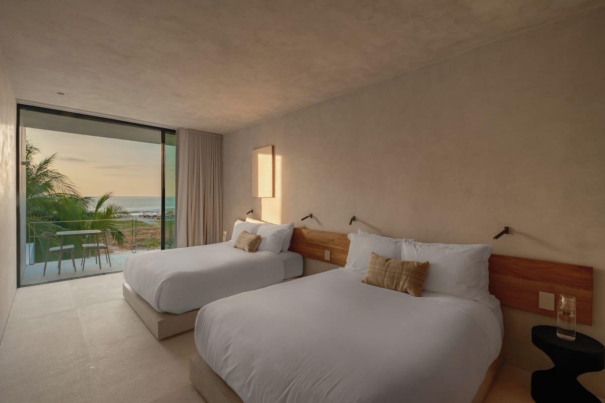 A hotel room with two beds, a large window with a view of the beach, and a small table with chairs.