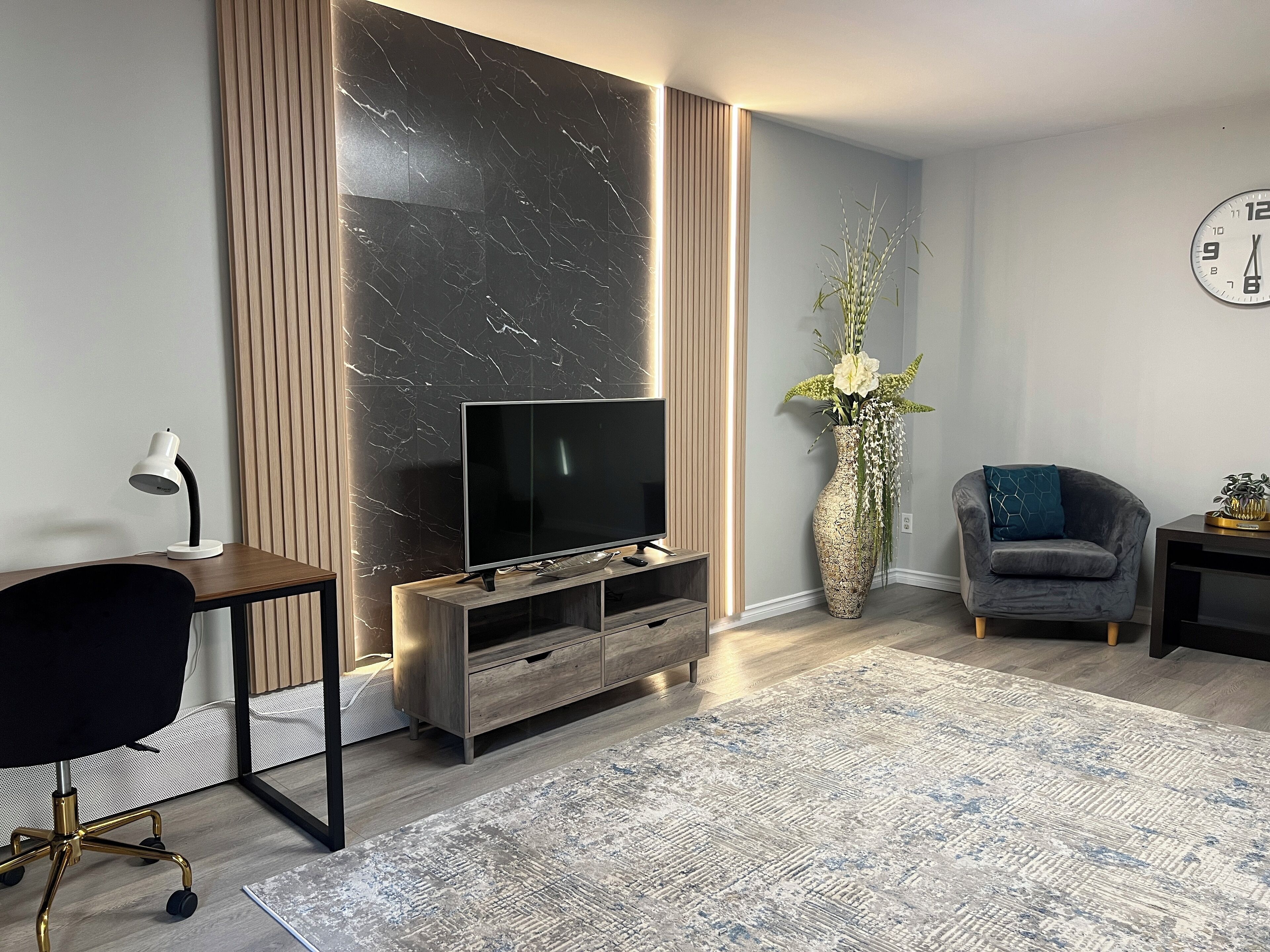 Un salon moderne avec un téléviseur à écran plat posé sur un meuble en bois, un fauteuil de bureau noir, un vase décoratif avec des fleurs, un fauteuil gris et une horloge au mur.