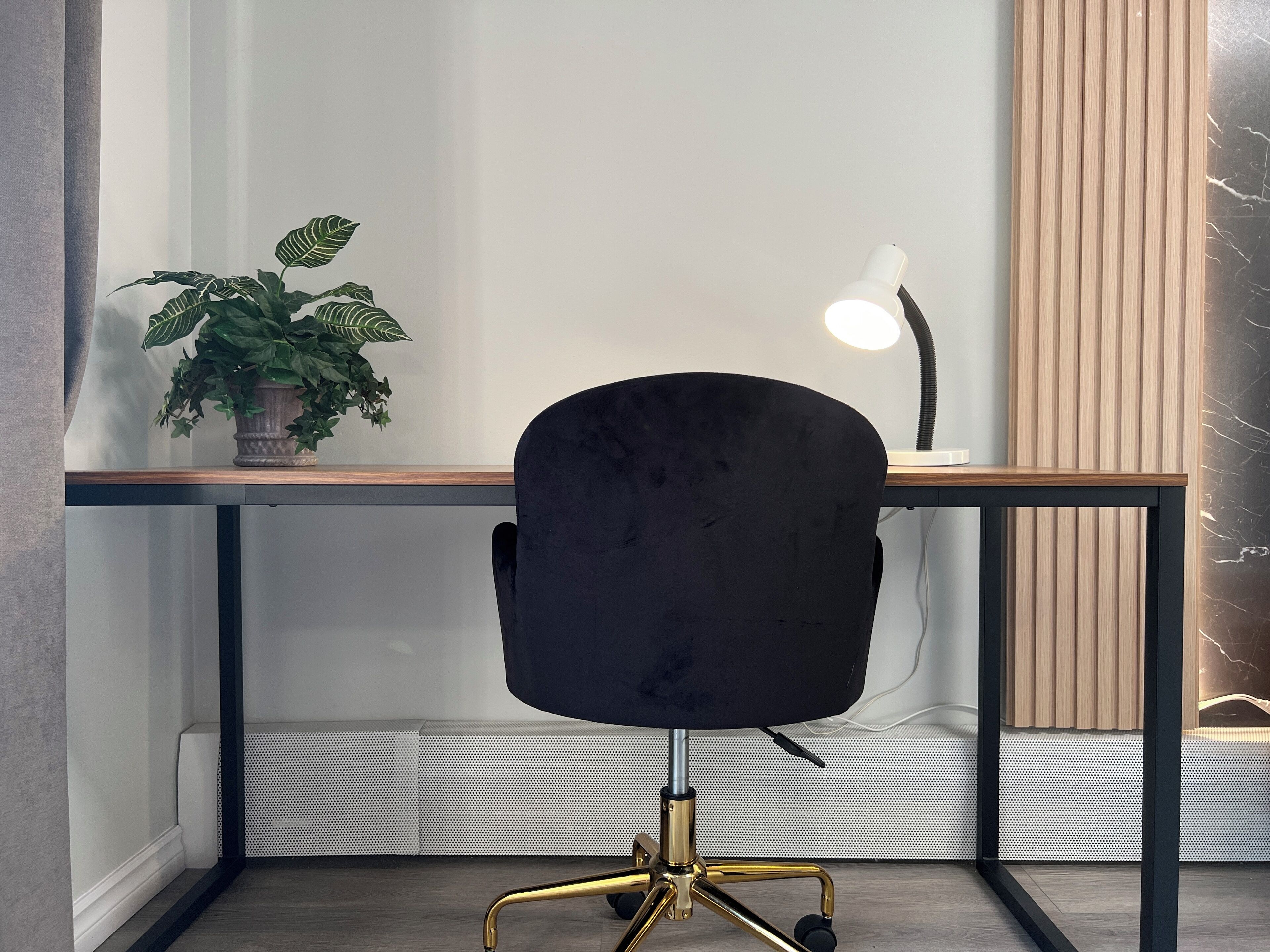 Una silla de oficina moderna con asiento negro y patas doradas, ubicada frente a un escritorio de madera con una planta en maceta y una lámpara de escritorio.