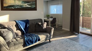 Un salón con un sofá gris, una manta azul y un cuadro de una cascada. Hay una mesa de comedor con sillas y una ventana con cortinas.