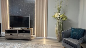 Una sala de estar moderna con un televisor de pantalla plana sobre un mueble de madera, un sofá gris con un cojín estampado y un jarrón alto con flores blancas.