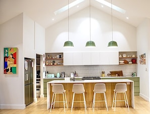 A modern kitchen with a central island, bar stools, pendant lights, and a refrigerator. - Luxury Coastal Gallery Retreat (Narrawallee)