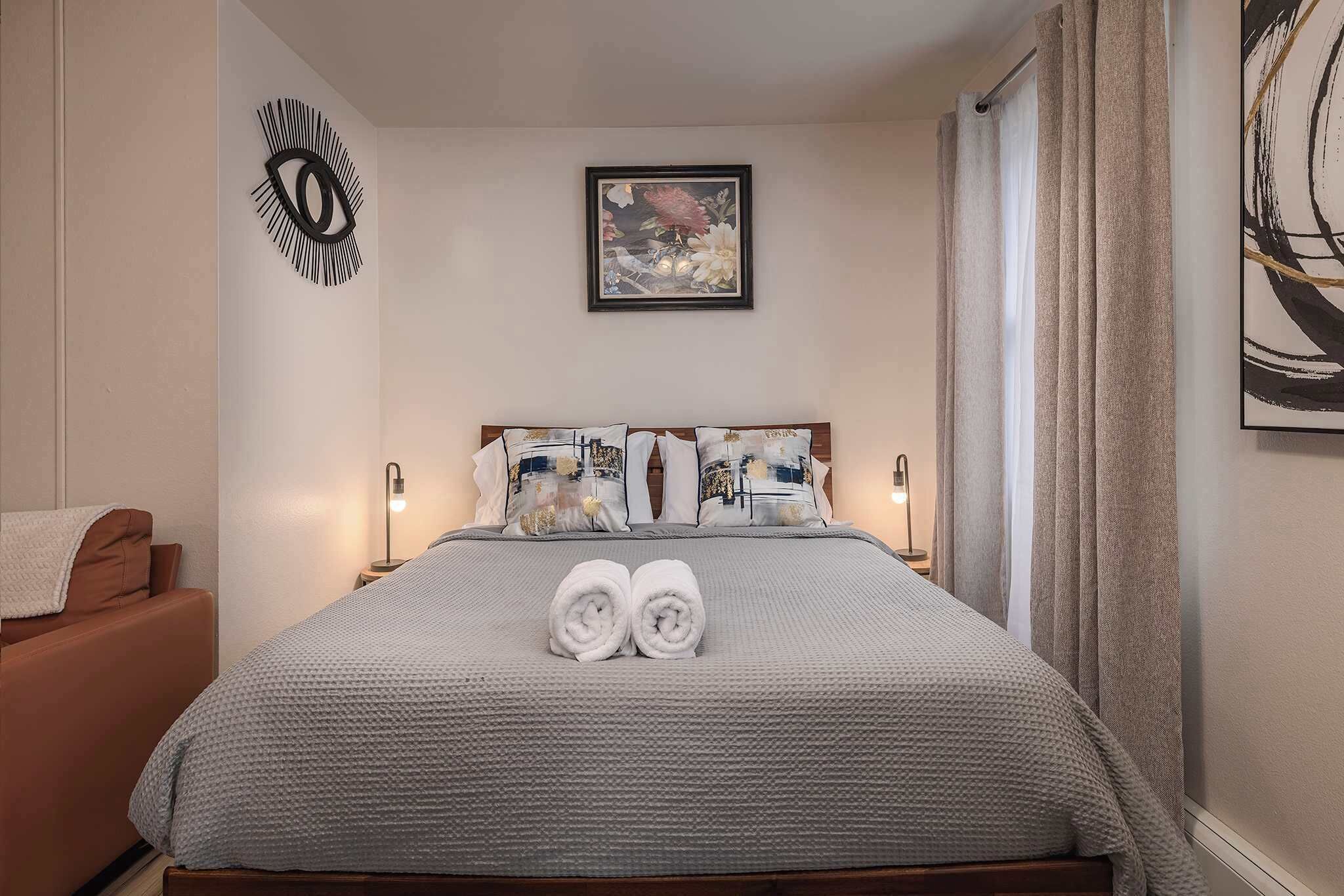 A neatly made bed with a grey textured bedspread, two rolled towels, and a headboard with decorative pillows.