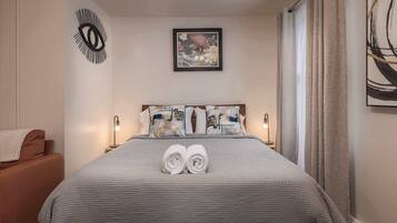 A neatly made bed with a grey textured bedspread, two rolled towels, and a headboard with decorative pillows.