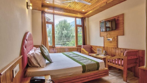 A wooden-paneled bedroom with a bed, a chair, a small table, a television mounted on the wall, and a window with a view of greenery. - The Veer Villa (Manali)