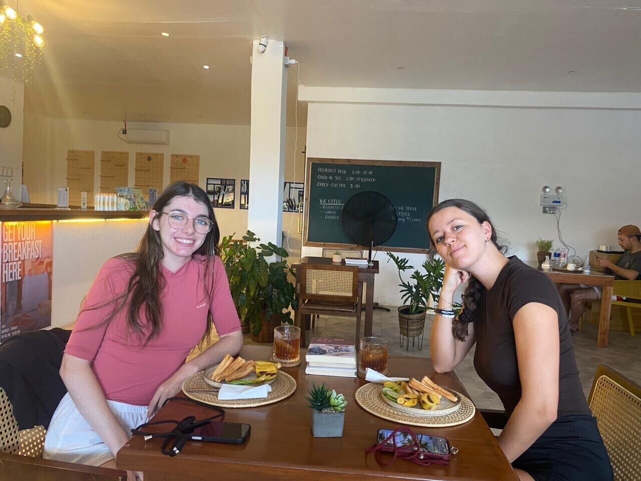 Two people sitting at a table with plates of food and drinks, in a room with a chalkboard menu and a plant in the background.