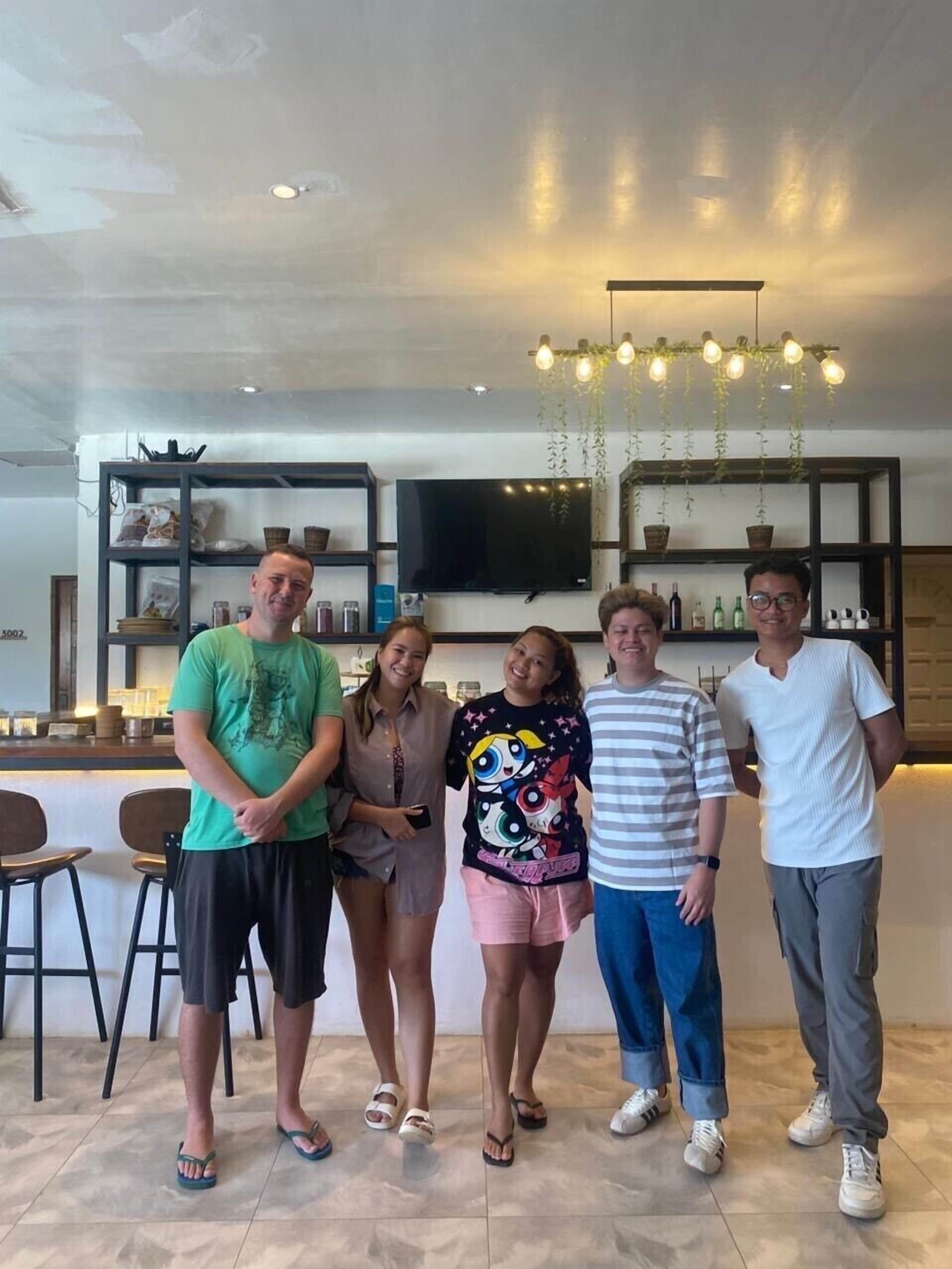 A group of people standing in a modern interior space with a bar area, shelves with bottles, and a flat-screen TV on the wall.