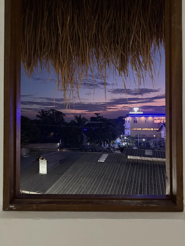 A view from a window with a wooden frame and a thatched roof, overlooking a rooftop area with a building labeled "DIAMONDRESTO" in the background.
