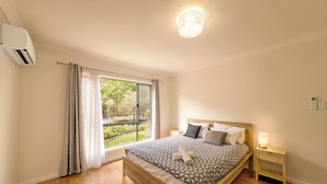 A bedroom with a large bed, a window with curtains, a chandelier, and a bedside table with a lamp.