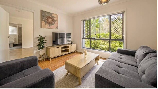 A modern living room with a grey sofa, a wooden coffee table, and a TV on a wooden stand. There is a large window with a view of greenery outside.
