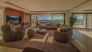 Ein modernes Wohnzimmer mit großem Fernseher, bequemen Sitzmöglichkeiten, einem Couchtisch und Blick ins Freie.