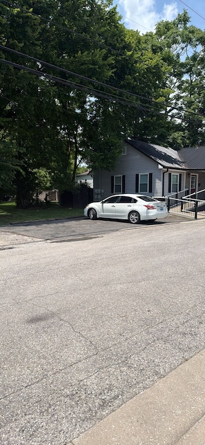 A residential street with a white car parked on the side, a house with a metal roof, and trees in the background.