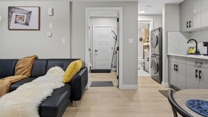 A modern living room with a black sofa, a white furry throw, and a yellow pillow. A hallway leads to a kitchen with a washing machine and cabinets. - Guest Suite in Edmonton(King bed & Driveway parking),Separate Entrance & Heating (Edmonton)