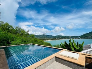 Un ciel d’un bleu limpide, une piscine dont le bord est recouvert de carreaux, et une vue sur une plage et des montagnes en arrière-plan.