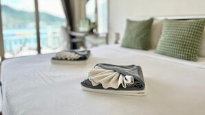 Een net opgemaakt bed met een opgevouwen handdoek en een paar schoenen erop, een groen kussen, en uitzicht op zee door grote ramen.