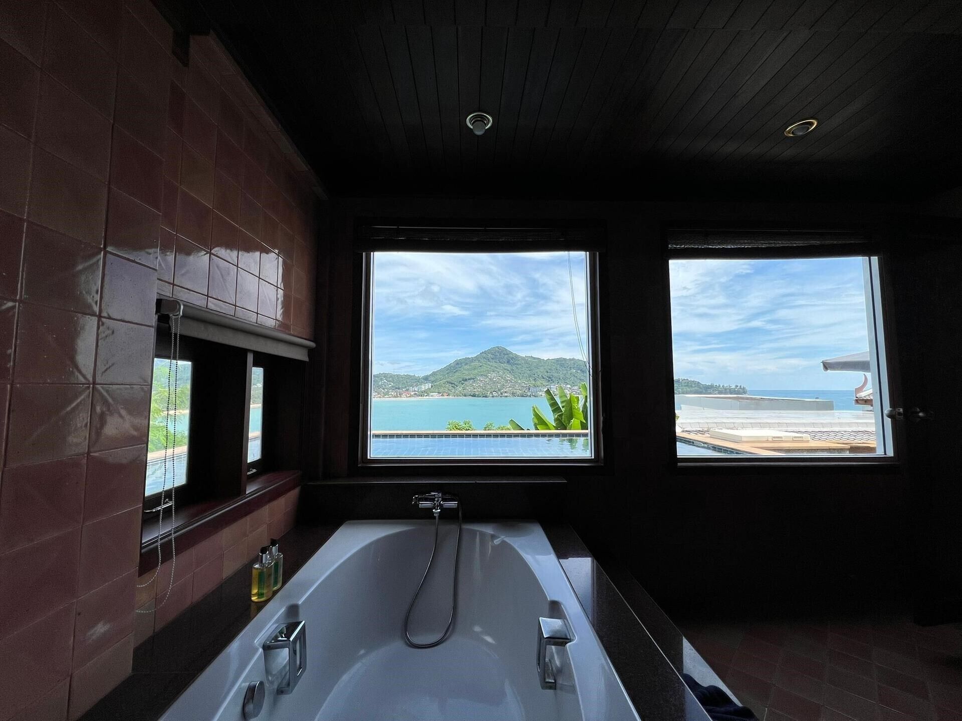 A modern bathroom with a large bathtub, two windows offering views of a coastal landscape, and a tiled wall.