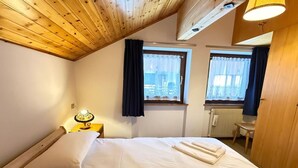 Ein Schlafzimmer mit Holzboden, zwei Fenstern mit Vorhängen, einem Bett mit weißer Bettwäsche, einer Nachttischlampe und einem Heizkörper.