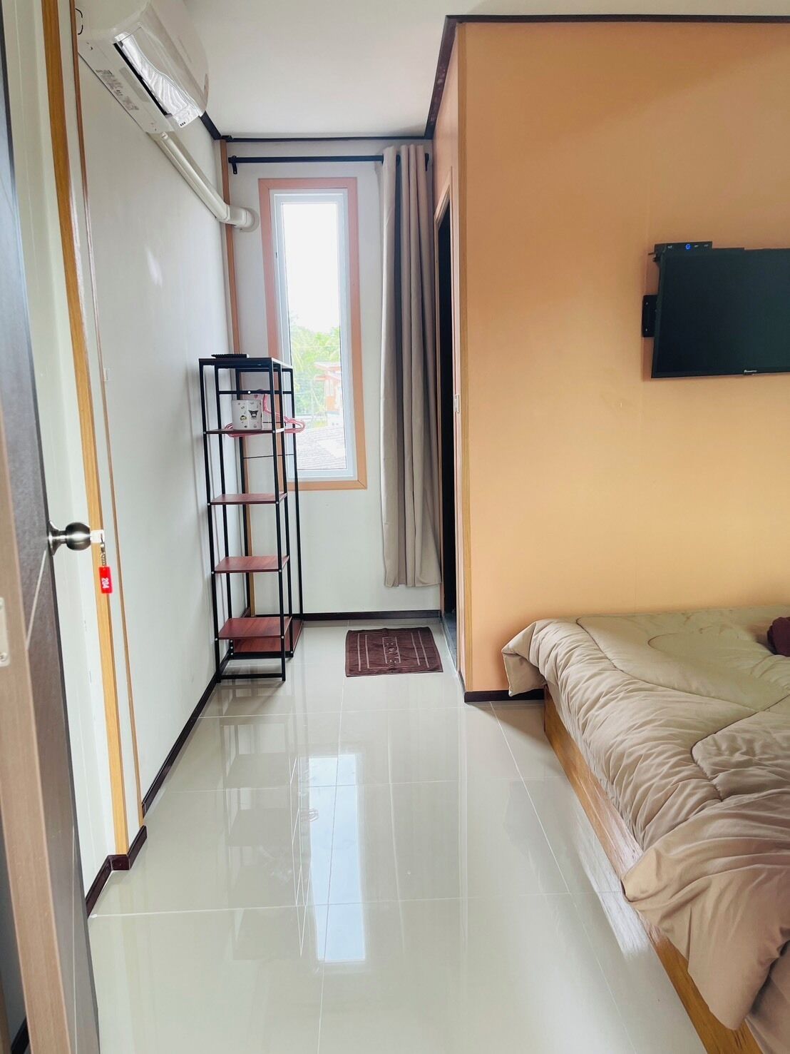 A hotel room with a bed, a TV on the wall, a shelf unit, and a door with an air conditioning unit.