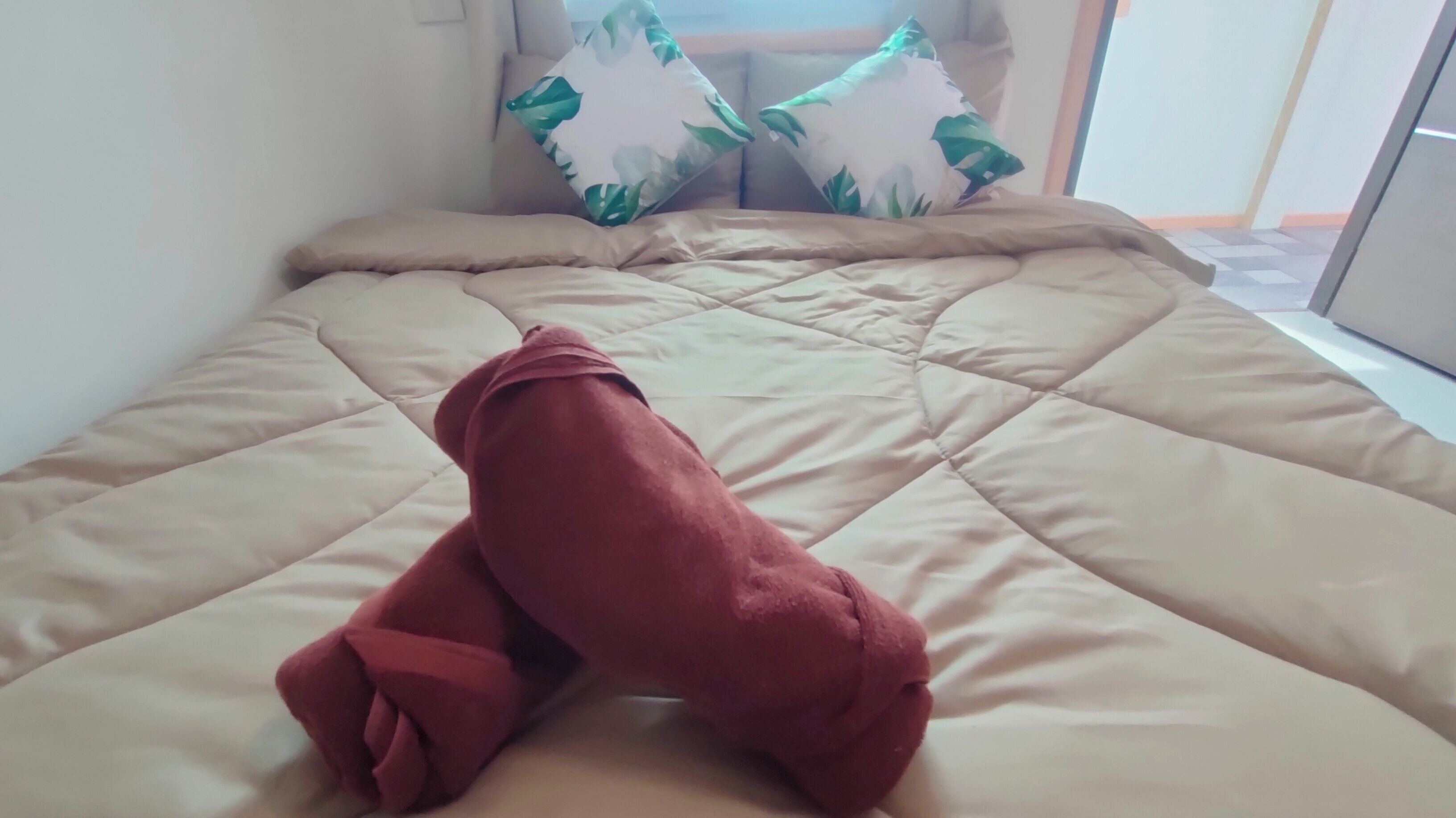 Een bed met een beige gewatteerde spreideken en een rode deken aan het voeteneinde.