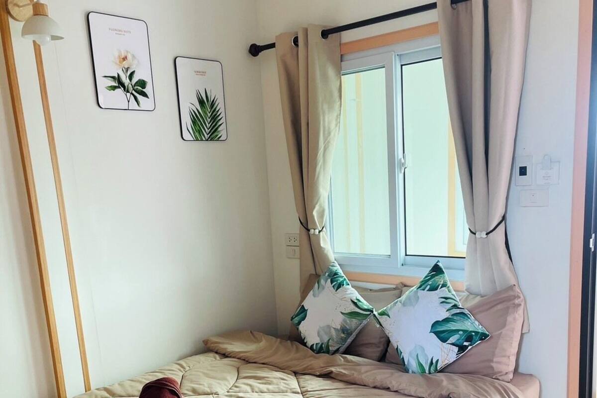 Una cama individual con colcha color beige, cabecera de madera y ventana con cortinas. Se observan dos lámparas de pared y dos láminas enmarcadas con motivos botánicos.