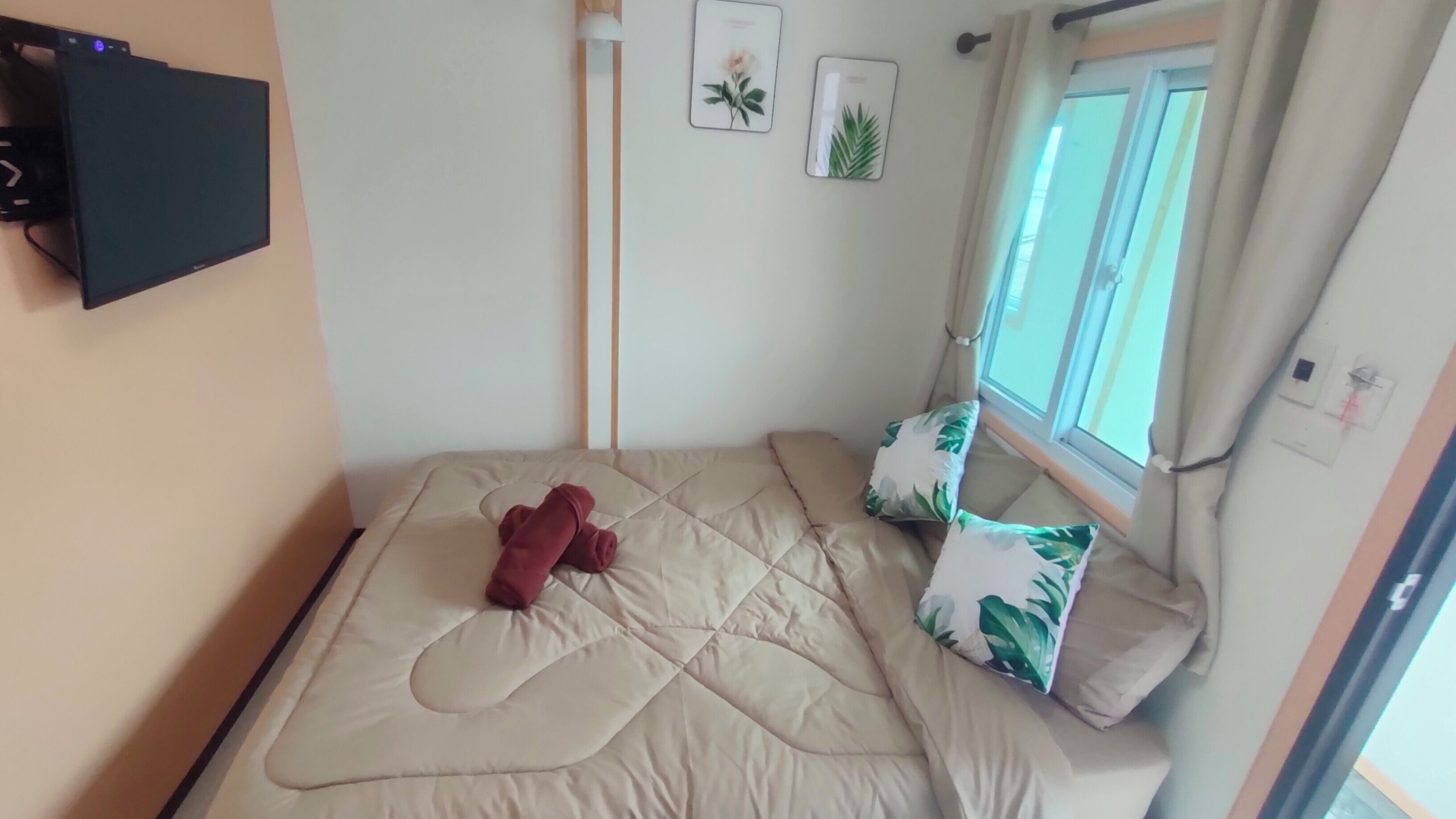 A small, neatly made bed with a beige quilt, two pillows with a green leaf pattern, and a brown item on the bed.
