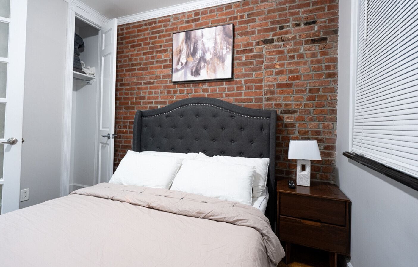 A bedroom with a brick wall, a bed with a grey upholstered headboard, white bedding, a nightstand with a lamp, and a framed picture on the wall.