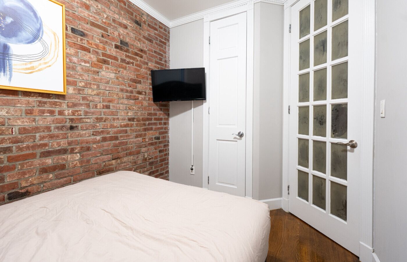 A bedroom with a brick wall, a flat-screen TV mounted above a white door, and a large window with frosted glass panels.