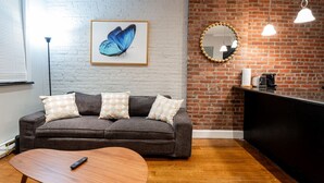 Una sala moderna con un sofá gris, una mesa de centro de madera, una obra de arte con una mariposa enmarcada, un espejo redondo y una pared de ladrillo.