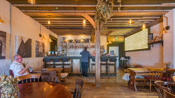 A restaurant interior with wooden tables and chairs, a bar area with a bartender, and a brick wall with framed pictures. - Pilgrim -  a holiday let that sleeps 2 guests  in 1 bedroom (Tenterden)
