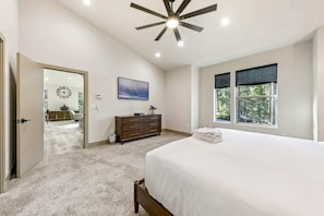 A bedroom with a large bed, a ceiling fan, a window with blinds, a dresser, and a painting on the wall.