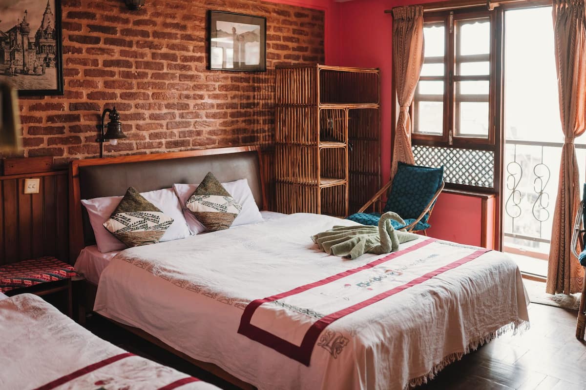Een kamer met een bakstenen muur, een bed met een witte spreideken en kussens, een rieten stoel en een raam met gordijnen.