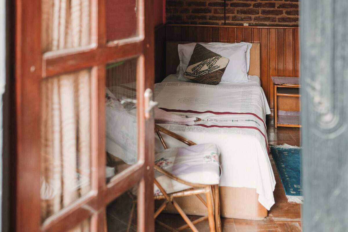 Een kamer met een bed, een houten stoel, een klein tafeltje en een bakstenen muur.