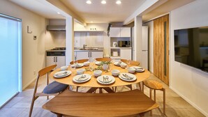 Un comedor moderno con una mesa redonda de madera dispuesta para seis personas, rodeada de sillas. Se ve una cocina al fondo con armarios blancos y una puerta de madera.