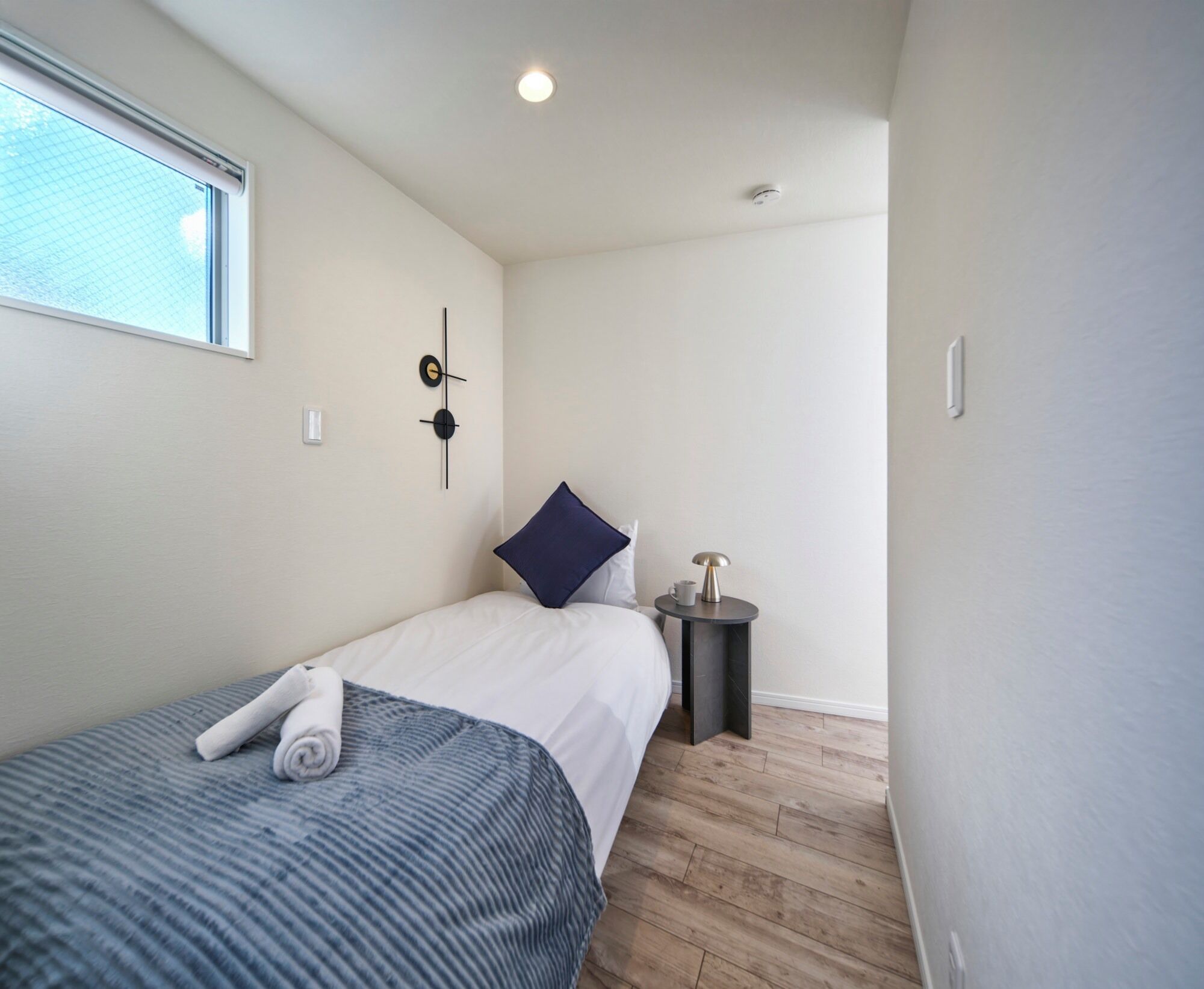 A bedroom with a bed, a small bedside table, a wall clock, and a window.