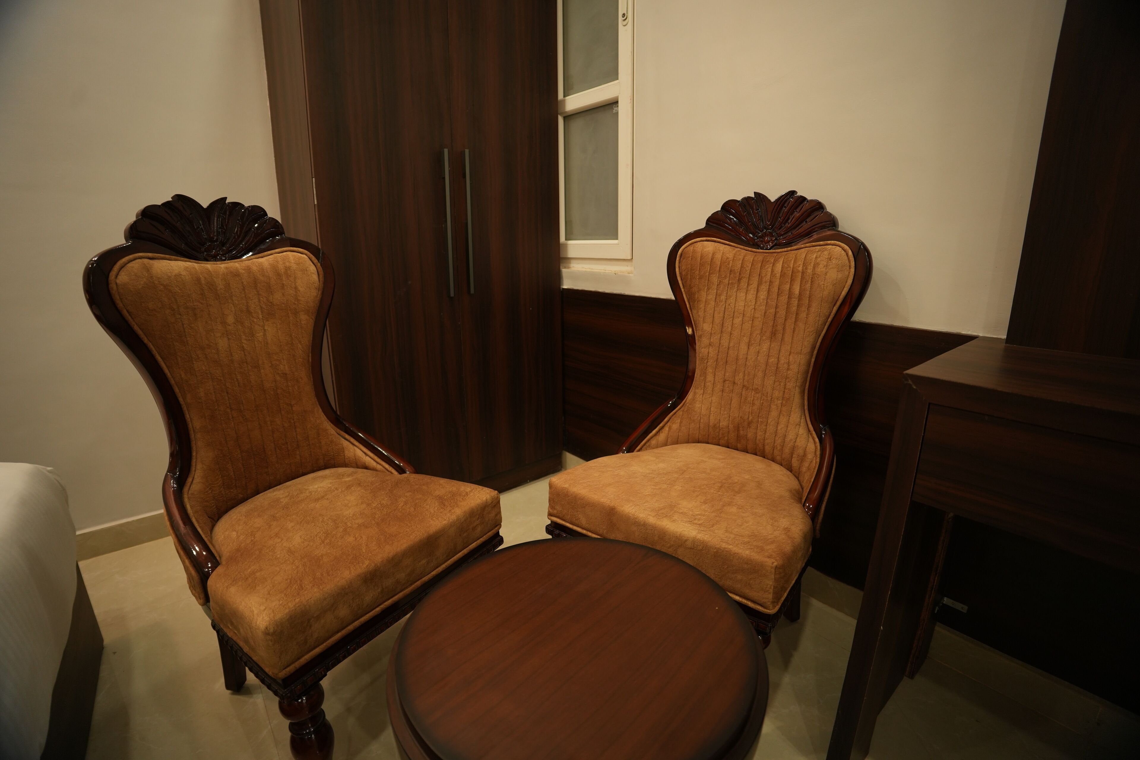 Two upholstered chairs with wooden frames and a round table in a room with a wooden wardrobe and a window.