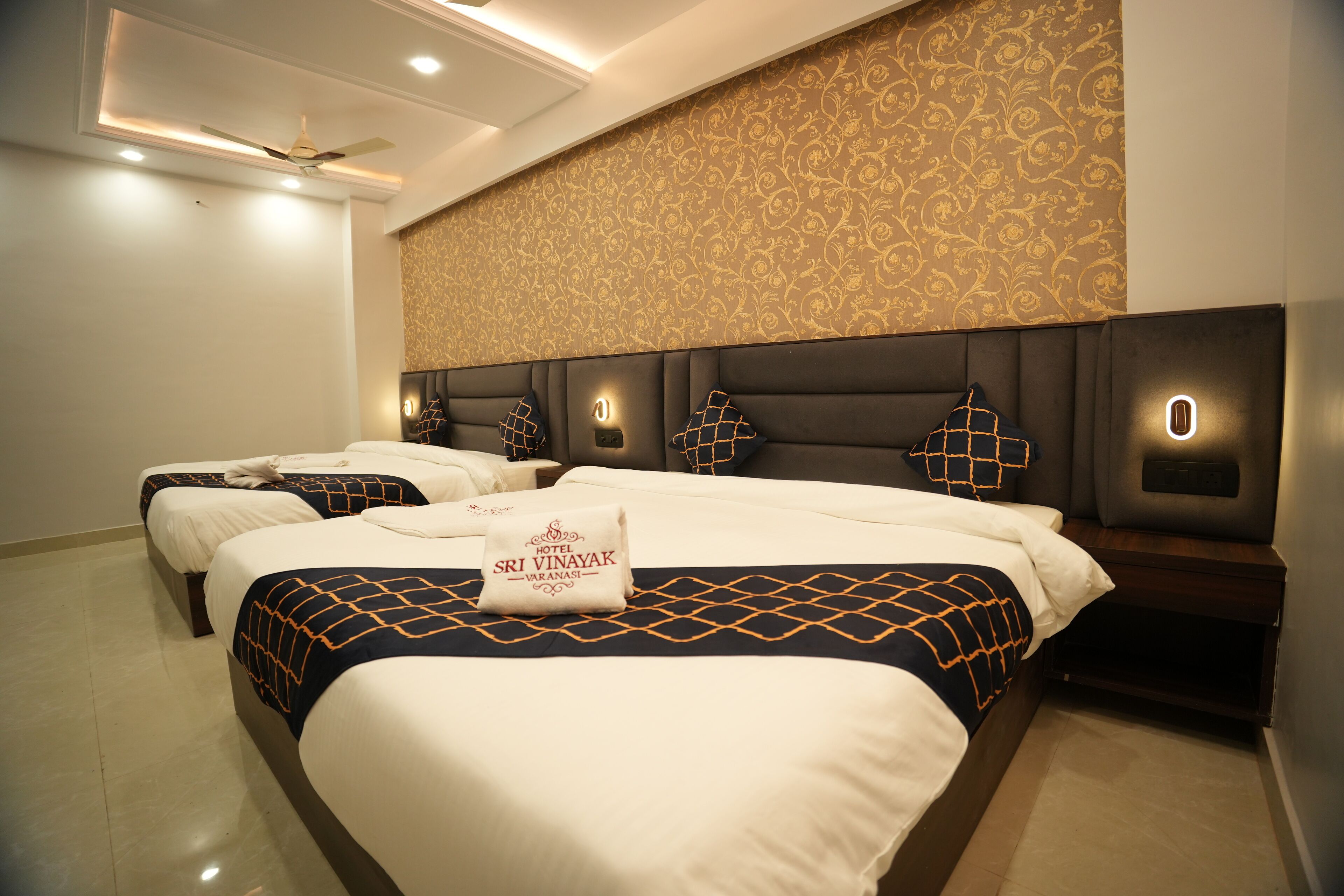A hotel room with two beds, each with a headboard, a ceiling fan, and wall-mounted lighting.