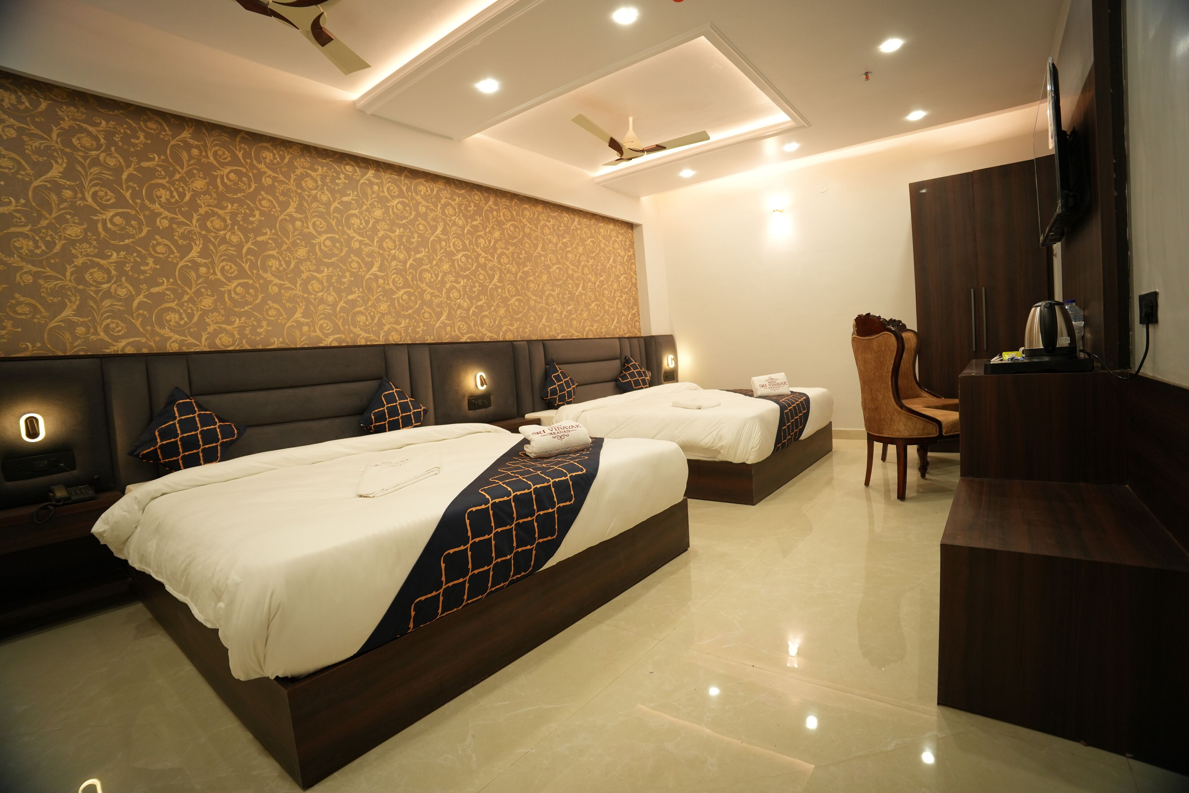 A hotel room with two beds, a desk, a chair, a TV, and a ceiling fan.