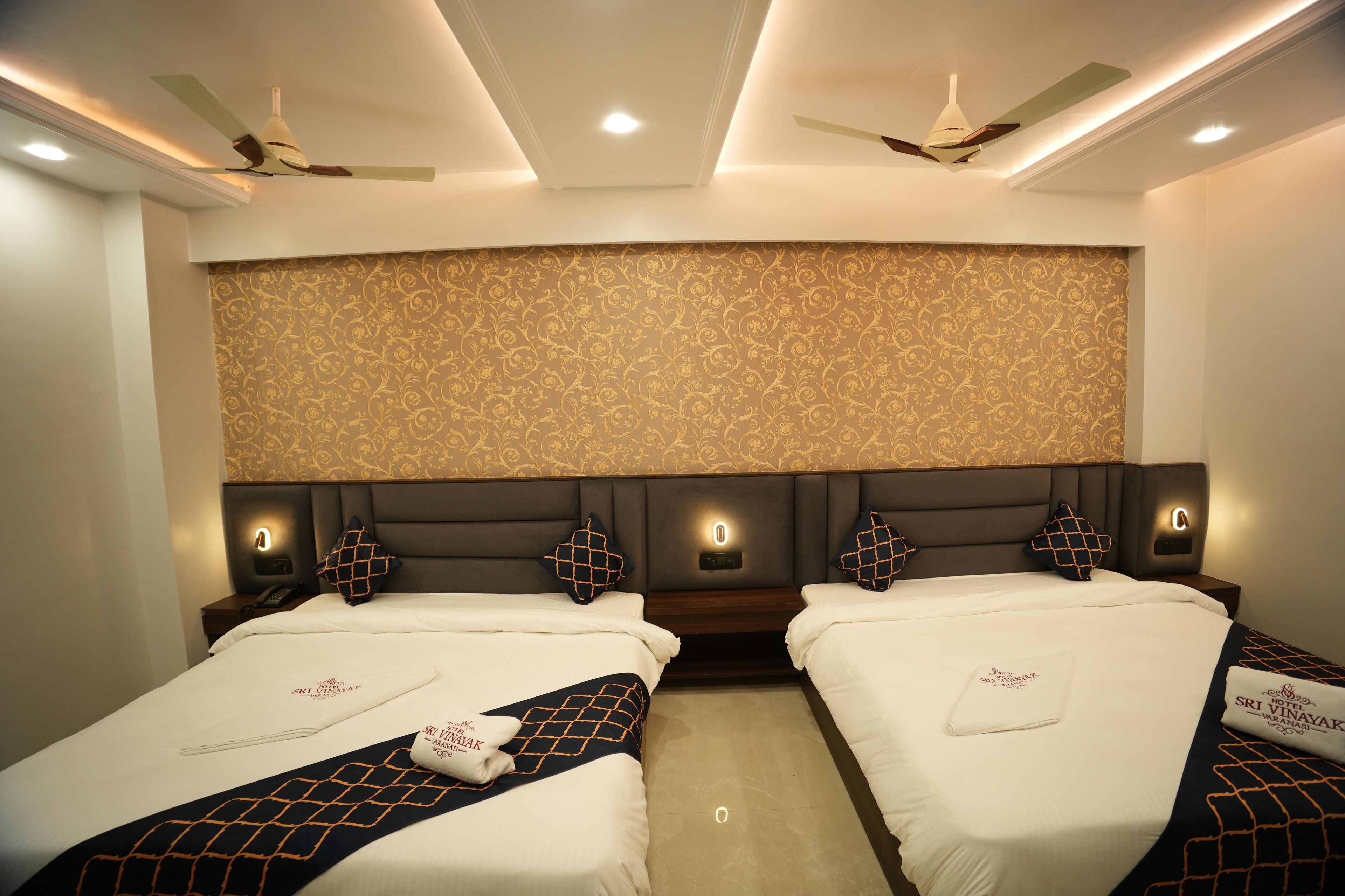 A hotel room with two beds, a patterned wallpaper, ceiling fans, and wall-mounted lights.