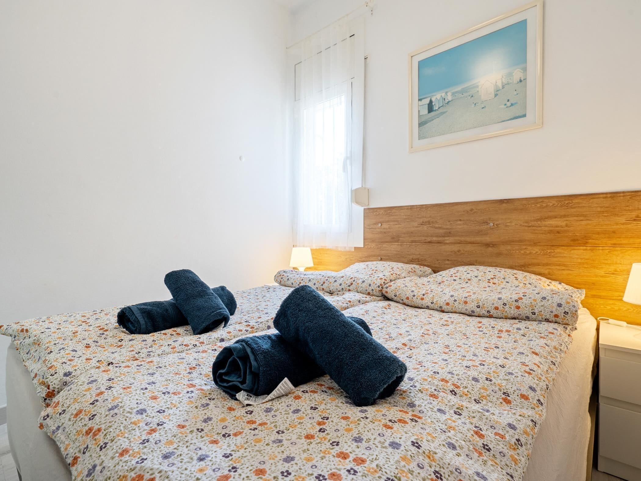Hotellihuone, jossa on sänky, yöpöytä, lamppu, ikkuna verhoilla ja seinällä oleva kehystetty kuva.