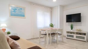 Ein modernes Wohnzimmer mit einer Couch, einem Esstisch mit Stühlen, einem an der Wand montierten Fernseher und einem Fenster mit Vorhängen.