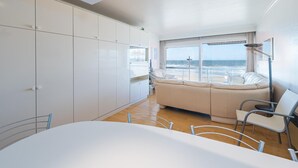 Quarto moderno com uma grande janela com vista para a praia, um sofá, uma mesa de jantar e armários brancos.