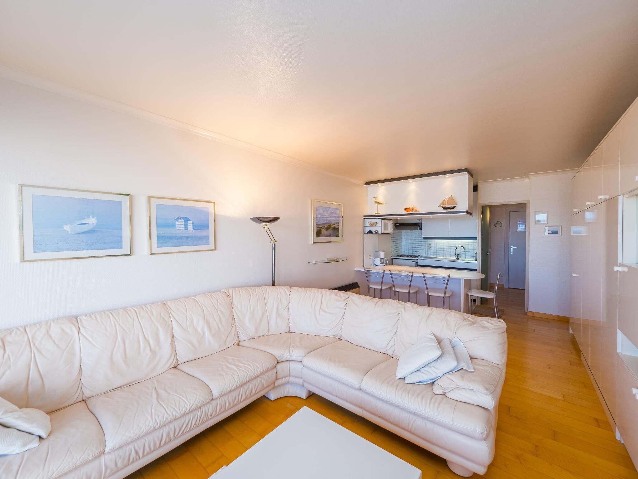 Een moderne woonkamer met een grote, beige L-vormige bank, een klein keukentje met een magnetron en een fornuis, en een eethoek met een barkruk.