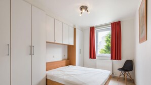 A bedroom with a bed, white cabinets, a window with red curtains, and a radiator. - Cosy flat close to the beach (Koksijde)