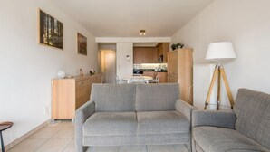 A modern living room with a grey sofa, a dining area with a white table and chairs, and a kitchen in the background. - Cosy flat close to the beach (Koksijde)