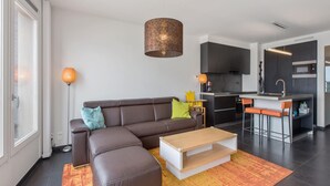 Een moderne woonkamer met een hoekbank, een houten salontafel en een keuken met donkere kastjes en een bargedeelte.