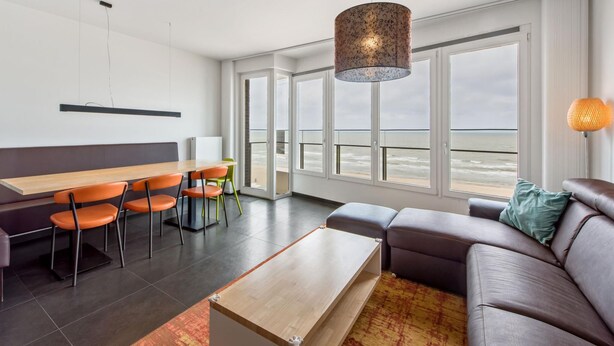 Een moderne woonkamer met een groot raam met uitzicht op zee, een bank, een eettafel met stoelen en een hanglamp.