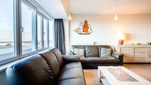 Una sala de estar moderna con un sofá de cuero, una mesa de centro con copas de vino, un adorno náutico en la pared y vistas a la playa a través de amplias ventanas.